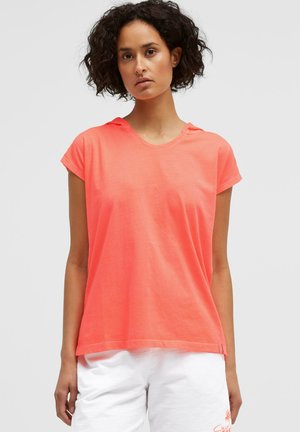 Woman with curly hair wearing coral short-sleeve hooded top and white shorts against a plain light background.
