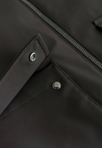 Gros plan sur un tissu noir avec un bouton-pression en métal et une fermeture éclair, mettant en valeur les coutures et les détails de la poche.