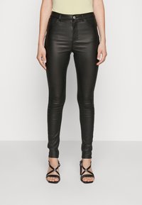 Jean en similicuir noir à coupe skinny, avec une taille haute et des coutures décoratives, associé à des sandales à talons noires.