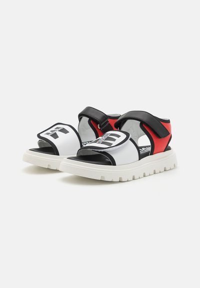 Sandalias abiertas en negro y blanco con una correa de Velcro, detalles en rojo y una suela blanca gruesa con textura ranurada.