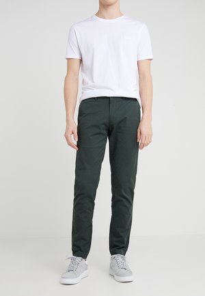 Mężczyzna ubrany w gładką białą koszulkę z krótkim rękawem, ciemnozielone spodnie o dopasowanym kroju oraz jasnoszare trampki, stojący na białym tle.