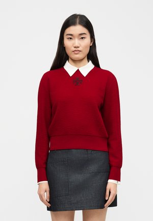 Femme aux longs cheveux noirs portant un pull rouge avec un col blanc et une jupe gris foncé, debout devant un fond clair uni.