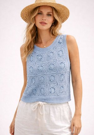 Femme aux cheveux blonds porte un chapeau de paille, un haut sans manches bleu crocheté et un pantalon blanc à cordon sur un fond clair uni.