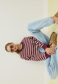 Maronenfarbener und cremegestreifter Pullover, hellblaue gerade Jeans, braune Slipper, übergroße braune Sonnenbrille, entspannte Pose auf dem Boden.