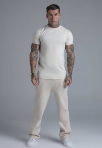 Cream short-sleeve t-shirt with a small logo on the chest, paired with light beige sweatpants featuring subtle branding, completed with white sneakers.
