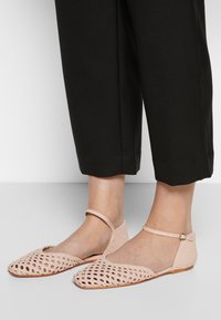 Chaussures plates tissées rose clair avec brides à la cheville, portées avec un pantalon noir court sur pieds nus, sur fond uni.