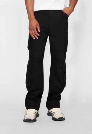 Férfi fekete többzsebes cargo nadrágban és fehér sportcipőben, egyik kezét a zsebében tartva, míg a másik oldalt nyugodtan pihenteti.