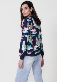 Floralna bomber jakna v temno modri barvi, s potiski v roza in zeleni tropski temi. Izdelana iz gladkega materiala, z rebrastimi manšetami in pasom.