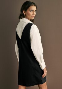 Robe en coton noire avec un dos en V, superposée à une chemise blanche à boutons. Présente une coupe décontractée et une texture lisse.