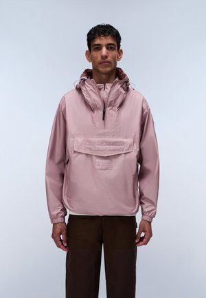 Homme portant un anorak à capuche rose clair avec poche avant et pantalon marron foncé, debout devant un fond clair uni.