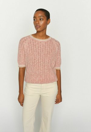 Rövid hajú nő rózsaszín bordás rövid ujjú pulóvert és krémszínű nadrágot viselve, egyszínű világos háttér előtt áll.