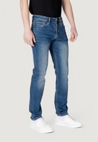 Jeans de mezclilla azul con un lavado desgastado, corte ajustado y diseño de cinco bolsillos, combinados con zapatillas blancas y una camiseta negra.