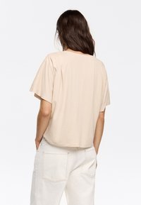 T-shirt beige a maniche corte con una vestibilità rilassata e scollatura rotonda, realizzato in tessuto morbido. Il retro presenta sottili dettagli di cucitura.