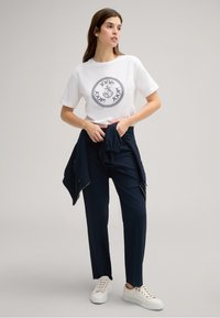 Witte katoenen t-shirt met een cirkelvormig zwart logo, gecombineerd met marineblauwe op maat gemaakte broek. Model houdt een marineblauwe jas vast en draagt witte sneakers.