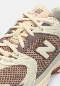 Running shoe with a mesh upper in light brown and cream. Features textured accents, a prominent "N" logo, and beige laces.