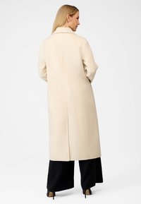 Beige lang frakke med krave, lavet af tekstureret stof, har en centerbag søm, og er parret med sorte vide bukser og højhælede sko.