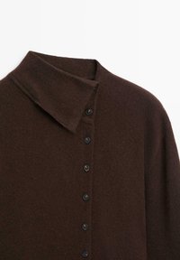 Cardigan lavorato a maglia marrone con colletto ripiegato, chiusura frontale con bottoni neri e una texture morbida. Focalizzato sulla semplicità e sul calore.