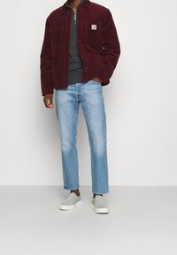 Hombre con una chaqueta de pana burdeos sobre un suéter negro de media cremallera, pantalones vaqueros de un azul claro y zapatillas ligeras de goma en color gris claro, de pie frente a un fondo blanco.