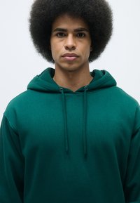 Felpa verde in cotone con cordini, caratterizzata da una texture morbida e una vestibilità rilassata. Il capo ha un design semplice senza motivi o grafiche.