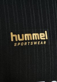 Golden gedrucktes „hummel SPORTSWEAR“-Logo auf strukturiertem schwarzem Stoff, wahrscheinlich Teil von Sport- oder Trainingskleidung.