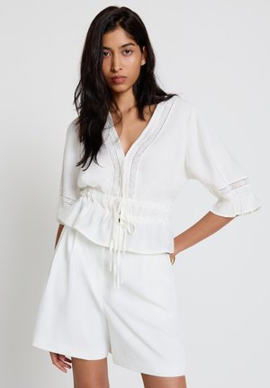 Jeune femme aux longs cheveux foncés portant un chemisier blanc à col en V avec des détails en dentelle et un short blanc taille haute, debout devant un fond uni.