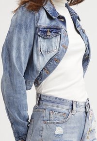 Giacca in denim azzurro chiaro a lunghezza cropped con colletto classico, chiusura frontale a bottone e due tasche sul petto. Presenta una finitura leggermente vissuta.
