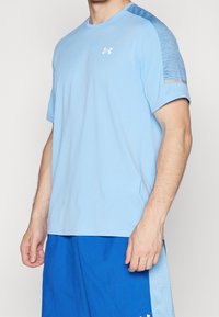 Ljusblå atletisk t-shirt med korta ärmar, med en texturerad blå panel på axeln och en vit logo på bröstet.