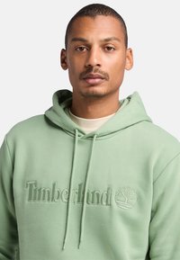 Felpa verde con cappuccio, con logo "Timberland" in rilievo e emblema ad albero. Tessuto liscio, design casual, cappuccio regolabile con cordino.