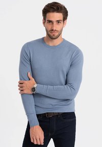 Lichtblauwe trui met lange mouwen van gebreid materiaal en een ronde halslijn. Geribbelde manchetten en tailleband. Model draagt donkere jeans en een zilveren horloge.