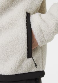 Chaqueta de forro polar blanca con una superficie texturizada, que presenta un ribete de tela negra en el bolsillo y el dobladillo, y un detalle de cordón en el bolsillo.