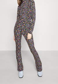 Eine Frau trägt einen enganliegenden, blumigen Jumpsuit mit langen Ärmeln und ausgestellt geschnittenen Beinen, kombiniert mit hellblauen Stiefeletten, und steht vor einem schlichten Hintergrund.