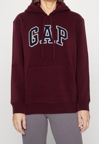 Sudadera con capucha color burdeos, con un bolsillo canguro frontal y un gran logo "GAP" en color marino. Presenta puños y dobladillo acanalados. Textura de tejido suave.