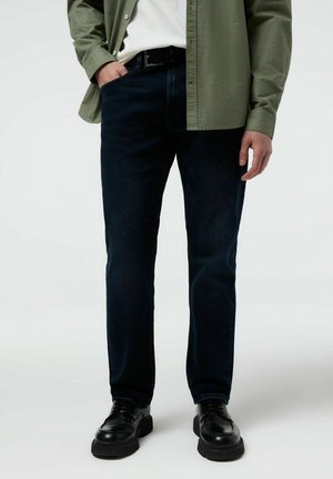 Mężczyzna w ciemnoniebieskich dżinsach, czarnych skórzanych butach, białej koszuli i rozpiętej oliwkowozielonej koszuli, stojący z jedną ręką w kieszeni.