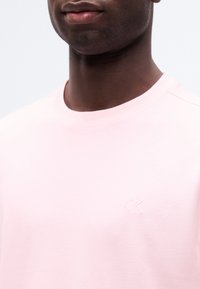 Κοντινό πλάνο ενός ατόμου που φοράει ένα ροζ ανοιχτό t-shirt με στρογγυλή λαιμόκοψη και μικρή κεντημένη λογότυπη "ck" στο στήθος.