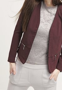 Bordeaux blazer med en tekstureret overflade, der har to frontlommer med lynlås, lagdelt over en grå blonde top og lysegrå bukser.