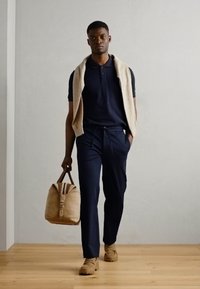 Marineblå polo skjorte kombinert med marineblå bukser, beige genser drapert over skuldrene, bærende en tannfarget duffelbag, og iført beige sko.
