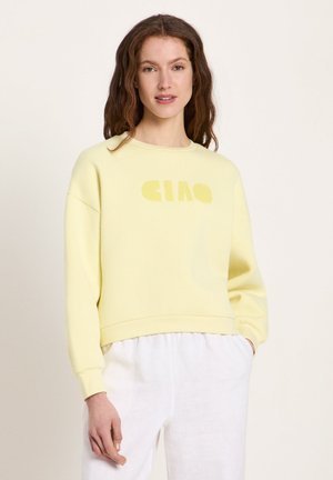 Frau mit langem braunem Haar, die einen hellgelben Pullover mit der Aufschrift "CIAO" und weiße Hosen trägt, steht vor einem hellen Hintergrund.
