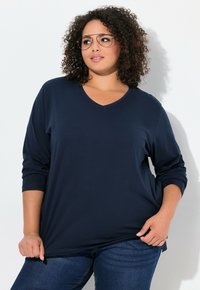 Navyblaues Langarm-V-Ausschnitt-Oberteil aus weichem Material, mit lockerer Passform, abgerundetem Saum und hochgekrempelten Ärmeln, kombiniert mit blauen Jeans.