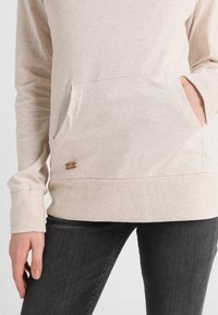 Beige långärmad sweatshirt med en framficka och ribbade ärmslut. Har en liten metall-logotag nära fållen. Mjukt tyg.