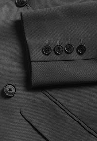 Must tume riie, millel on heledad triibud. Omab volditud varrukat, millel on neli musta nuppu ja logonupp all.