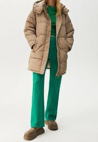Manteau beige matelassé avec capuche, doté de fermetures à boutons-pression. Porté sur un pull vert et un pantalon flare vert, complété par des chaussures épaisses marron.