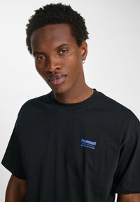 Zwarte katoenen T-shirt met een blauw "hummel" logo op de linkerborst. Ronde halslijn, korte mouwen en een relaxte pasvorm. Gladde textuur.