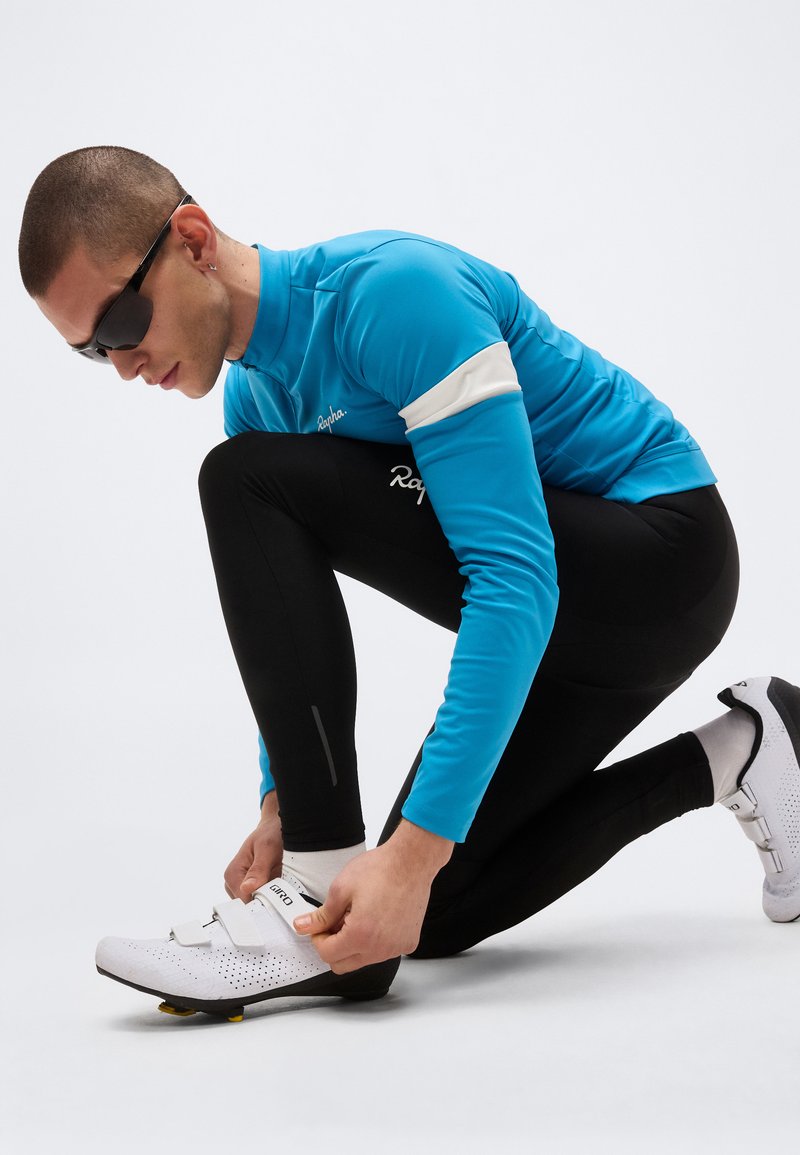 Athlete in blue cycling jacket and black leggings kneels to fasten white cycling shoe with cleats on plain background.