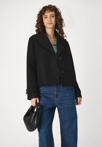 Black cropped jacket with wide collar and two buttons. Paired with blue wide-leg jeans and a small black handbag. Smooth fabric texture.