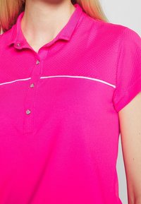 Světle růžové polo tričko s texturovanou tkaninou, se třemi knoflíky vpředu a bílým akcentním pruhem přes hruď.