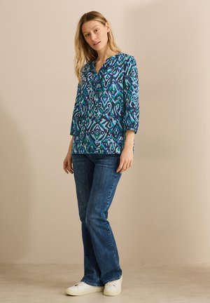 Multikleurige blouse met een blauw en groen abstract patroon, V-hals en driekwart mouwen, gecombineerd met donkere jeans en witte sneakers.