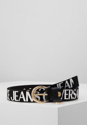 Zwarte stoffen riem met grote witte letters "VERSACE JEANS", gouden, afgeronde gesp en een kleine gouden klinknagel, opgerold weergegeven op een witte ondergrond.