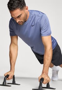 Μπλε αθλητική κοντομάνικη μπλούζα με μαύρο λογότυπο Nike, συνδυασμένη με μαύρο σορτς. Άτομο εκτελεί κάμψεις σε μαύρες λαβές μπροστά σε ανοιχτό φόντο.