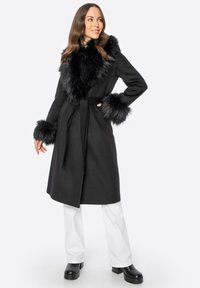 Schwarzer Mantel mit Kunstpelzkragen und -manschetten, tailliertem Bund, langen Ärmeln und knielangem Design, kombiniert mit weißen Hosen und klobigen schwarzen Schuhen.