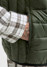 Zielona kamizelka z wypełnieniem o strukturalnym wzorze, z kieszenią na zamek błyskawiczny, w połączeniu z brązową i białą koszulą w kratę.